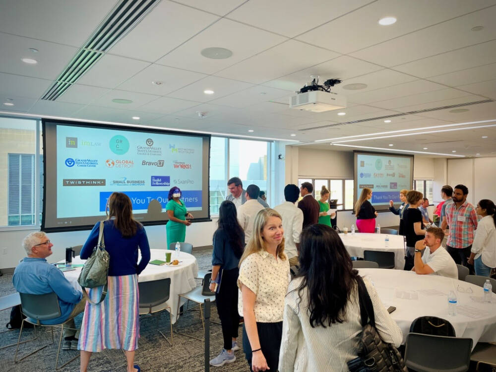 A group of people are networking in a bright, modern conference room with round tables and large windows. Two projection screens display a slide that thanks various partner organizations, and the room has a collaborative, casual atmosphere.